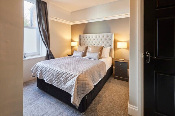 A bedroom with a large bed, two bedside tables, a dark door, and a window with curtains. - Host Stay Endeavour Sails (Whitby)