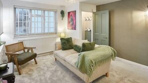 Een gezellige woonkamer met een bank, een houten stoel, een radiator, een raam met zonwering, een schilderij en een deur.