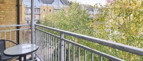Un balcon agrémenté d’une table et de chaises, entouré d’une rambarde en métal, donnant sur un quartier résidentiel avec des maisons et des arbres.