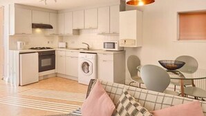 A compact kitchen with white cabinets, a microwave, a stove, and a washing machine. There is a dining area with a glass table and chairs, and a sofa with patterned cushions. - Host Stay King s Corner (Canterbury)