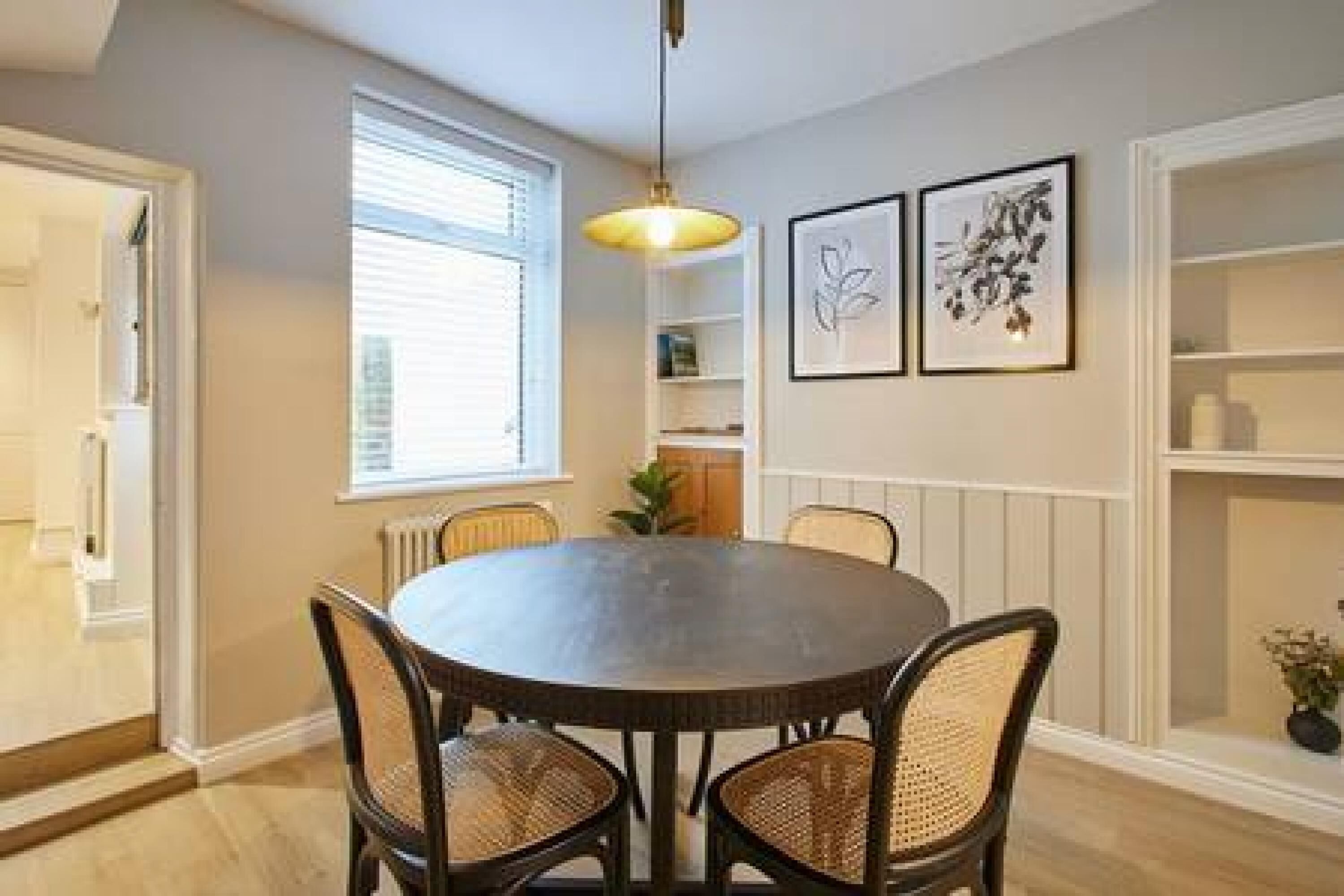 Uma área de refeições com uma mesa redonda, quatro cadeiras de vime, uma luminária pendente e quadros decorativos na parede.