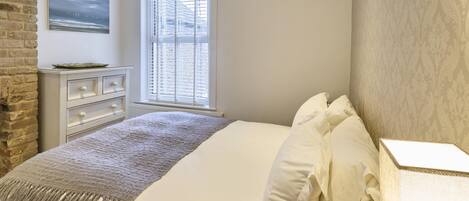 A bedroom with a bed, a dresser, a window with blinds, a wall-mounted painting, and a patterned wall.