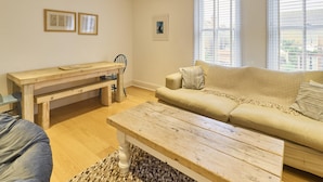 A living room with a wooden bench, a sofa, and a coffee table. There are framed pictures on the wall and a window with blinds. - Host Stay Sail Away (Whitstable)