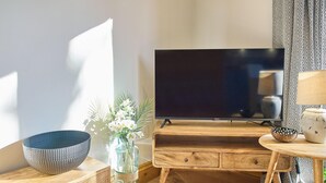 Ett mysigt vardagsrum med ett TV-bord i trä, en vas i glas med blommor, en blå skål och ett litet runt bord med en lampa.