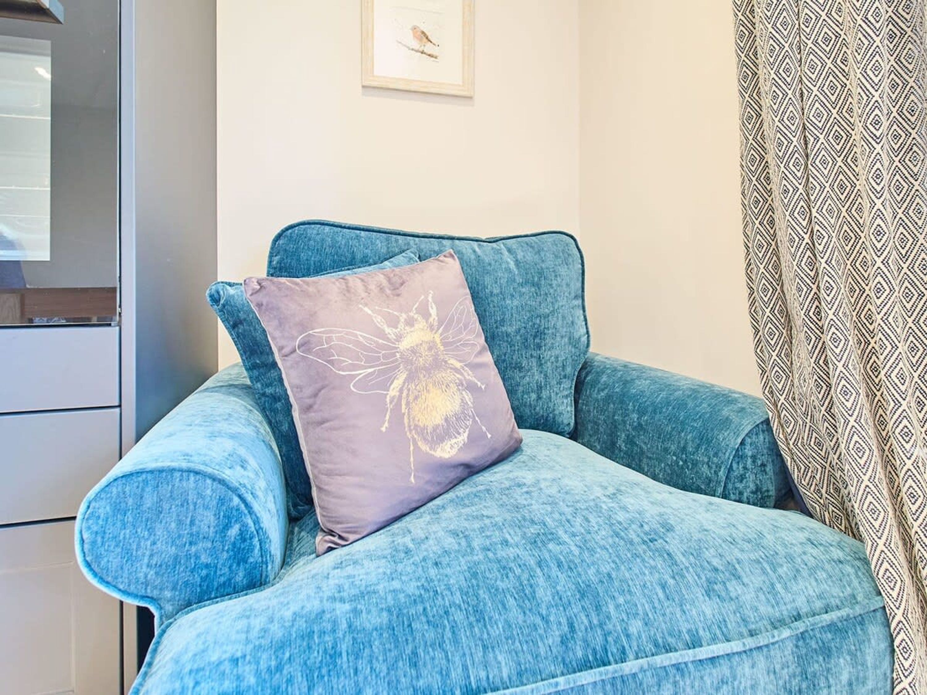 A blue armchair with a purple cushion featuring a bee design, positioned in a room with a white wall and a framed picture.