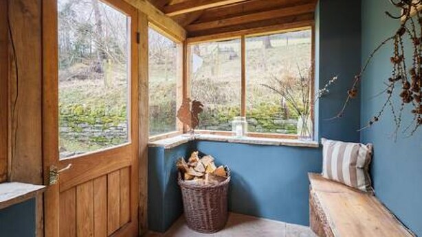 A cozy corner with a wooden bench, a wicker basket filled with firewood, and a window offering a view of the outdoors. - Host Stay Llewenau Draw Barn (Crickhowell)