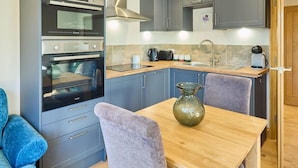 A compact kitchen with grey cabinets, a double oven, a sink, and a wooden dining table with a vase. - Host Stay Willow Barn (Ashford)