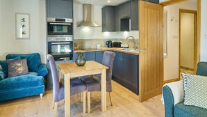 A compact kitchen with modern appliances, a dining area with a wooden table and chairs, and a blue sofa with a decorative pillow. - Host Stay Willow Barn (Ashford)