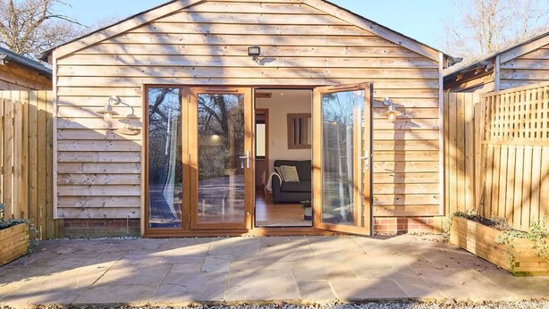 A wooden cabin with a large sliding glass door entrance, a small porch area, and a visible living room with a sofa and a coffee table. - Host Stay Willow Barn (Ashford)