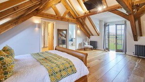 A bedroom with a sloped ceiling, wooden beams, a bed with a patterned blanket, a chair, and a view of the outdoors. - Host Stay Coed Barn (Brecon)
