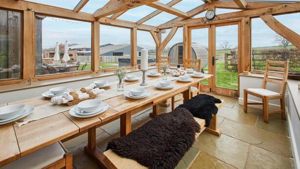 A dining area with a long wooden table set for a meal, large windows, and a view of the countryside. - Host Stay Coed Barn (Brecon)