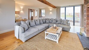 A modern living room with a large L-shaped grey sofa, a wooden coffee table, and a large window offering a view of the outdoors. - Host Stay Coed Barn (Brecon)
