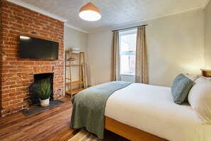 A bedroom with a brick fireplace, a bed with white bedding, a television mounted on the wall, and a window with curtains.