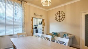 Un coin repas avec une longue table, des chaises blanches, une horloge au mur et une fenêtre avec des stores.