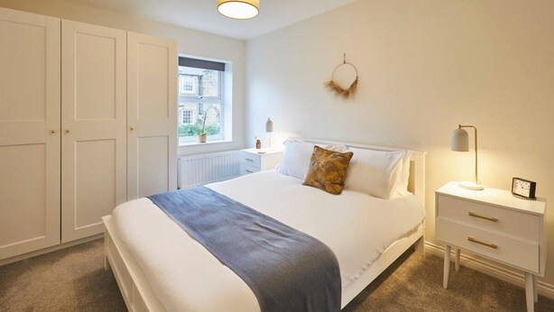 A bedroom with a large bed, a bedside table, a wardrobe, and a window with a view of buildings. - Host Stay Column Mews (Alnwick)