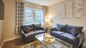 A living room with two grey sofas, a glass coffee table, and a lamp. There are three wall art pieces above the sofa. - Host Stay Column Mews (Alnwick)