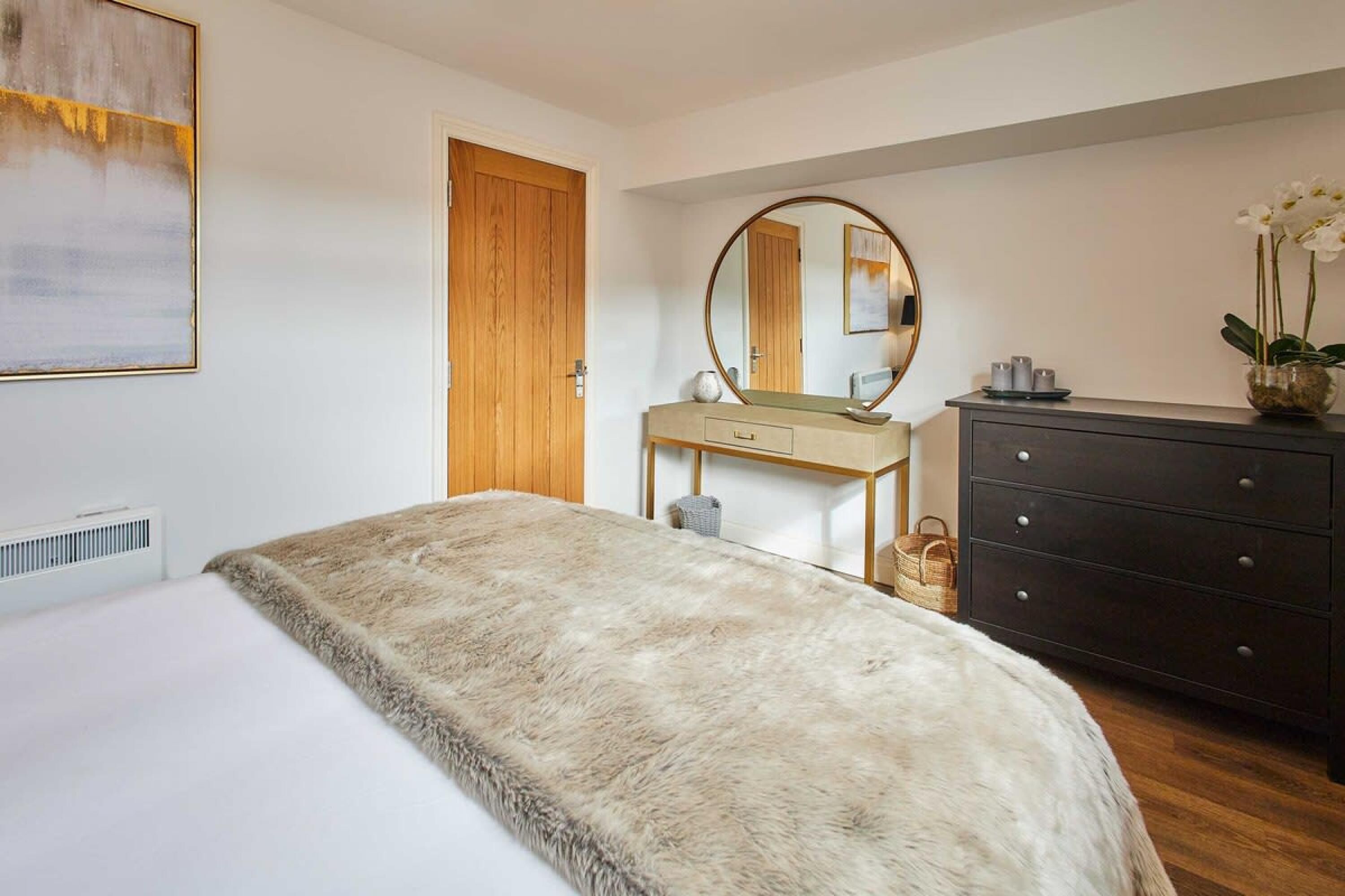 A bedroom with a bed, a wooden door, a dresser, a desk, and a mirror.