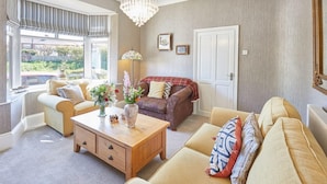 A living room with a beige sofa, a wooden coffee table, and a red plaid sofa. There are two windows with blinds, a chandelier, and a door. - Host Stay The Beach House (Redcar)