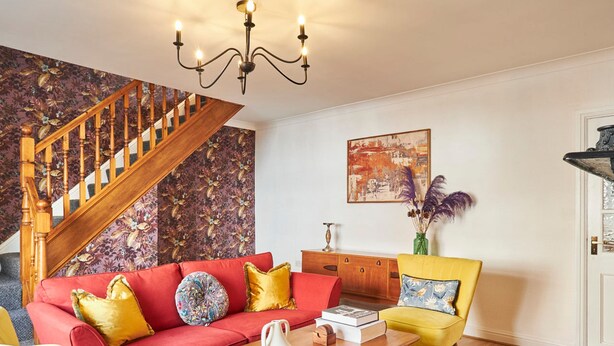 A living room with a red sofa, yellow chairs, a wooden coffee table, a chandelier, a staircase, and a floral wallpaper. - Host Stay Hideaway (Morpeth)