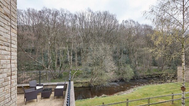 An outdoor patio with wicker furniture, a brick wall, and a view of a river and forest. - Host Stay 57 Woodland View (Holmfirth)
