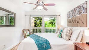 A bedroom with a bed, a ceiling fan, a window with a view of greenery, and a wall with a beach-themed artwork.