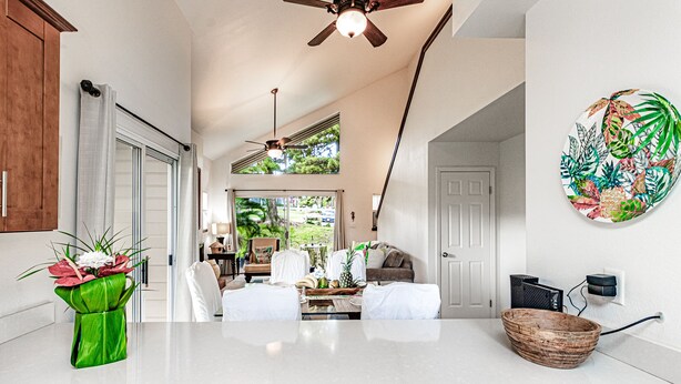 A spacious living area with a high ceiling, a ceiling fan, and a view of the outdoors. There is a dining area with a table and chairs, and a large decorative plant on the wall.