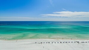 Une plage aux eaux turquoise et limpides, un rivage sablonneux, et quelques personnes ainsi que des chaises de plage.