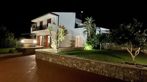 A modern two-story house with a stone retaining wall, a well-lit entrance, and a small garden with trees and shrubs. - Villa Lisa holiday home (Città del Mare-perla del Golfo)