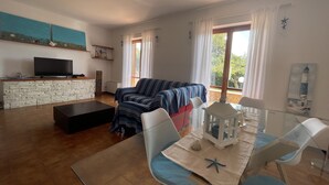 A living room with a stone fireplace, a sofa, a TV, and a dining area with chairs and a table. - Villa Lisa holiday home (Città del Mare-perla del Golfo)
