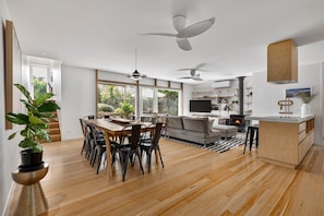 A spacious living area with a wooden dining table, black chairs, a grey sofa, and a TV. There are ceiling fans, a fireplace, and a large window offering a view of the outdoors.