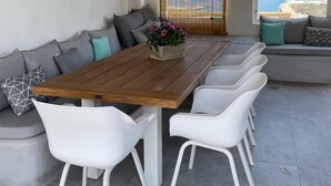 Un coin repas avec une table en bois, des chaises blanches, un canapé avec des coussins et une vue sur la mer grâce à de grandes fenêtres.