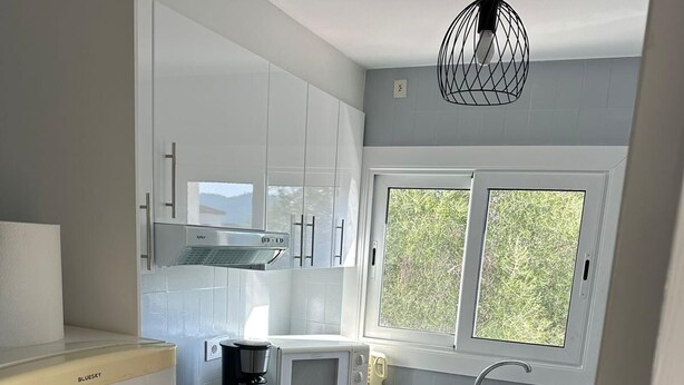 A compact kitchen with white cabinets, a built-in oven, a microwave, and a coffee maker. There is a window with a view of greenery outside.