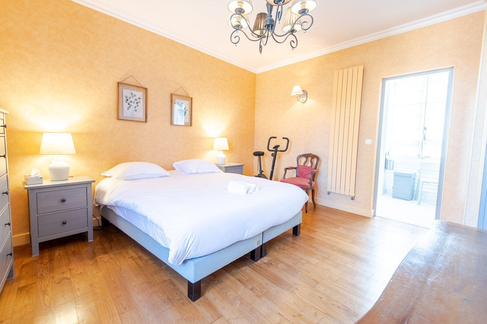 A bedroom with a large bed, two bedside tables with lamps, a chair, a radiator, and a chandelier.