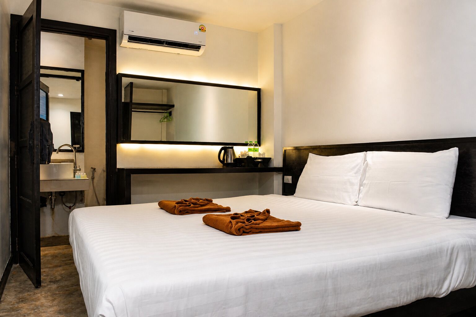 A hotel room with a bed, a bedside table, a mirror, an air conditioning unit, and a bathroom visible through an open door.