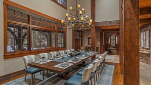 Une grande salle à manger avec une longue table en bois dressée pour un repas, bordée de grandes fenêtres offrant une vue sur les arbres et sur un ours à l’extérieur.