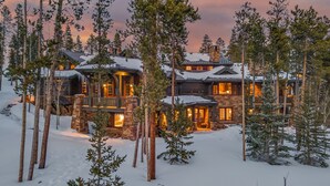 Un grand chalet à plusieurs étages, construit en pierre et en bois, entouré de neige et de pins.