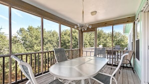 A covered outdoor deck with a glass-top table and chairs, surrounded by a wooden railing, overlooking a wooded area.