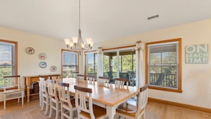 A dining room with a wooden table and chairs, large windows with blinds, and a chandelier.