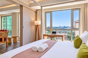 A hotel room with a large bed, a desk, a chair, and a view of the city through large windows. - Capital M Apartments by YourHost (Nairobi)