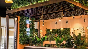 Interior de restaurante com design moderno, apresentando uma parede verde com plantas, mesas de madeira e assentos estofados em preto.