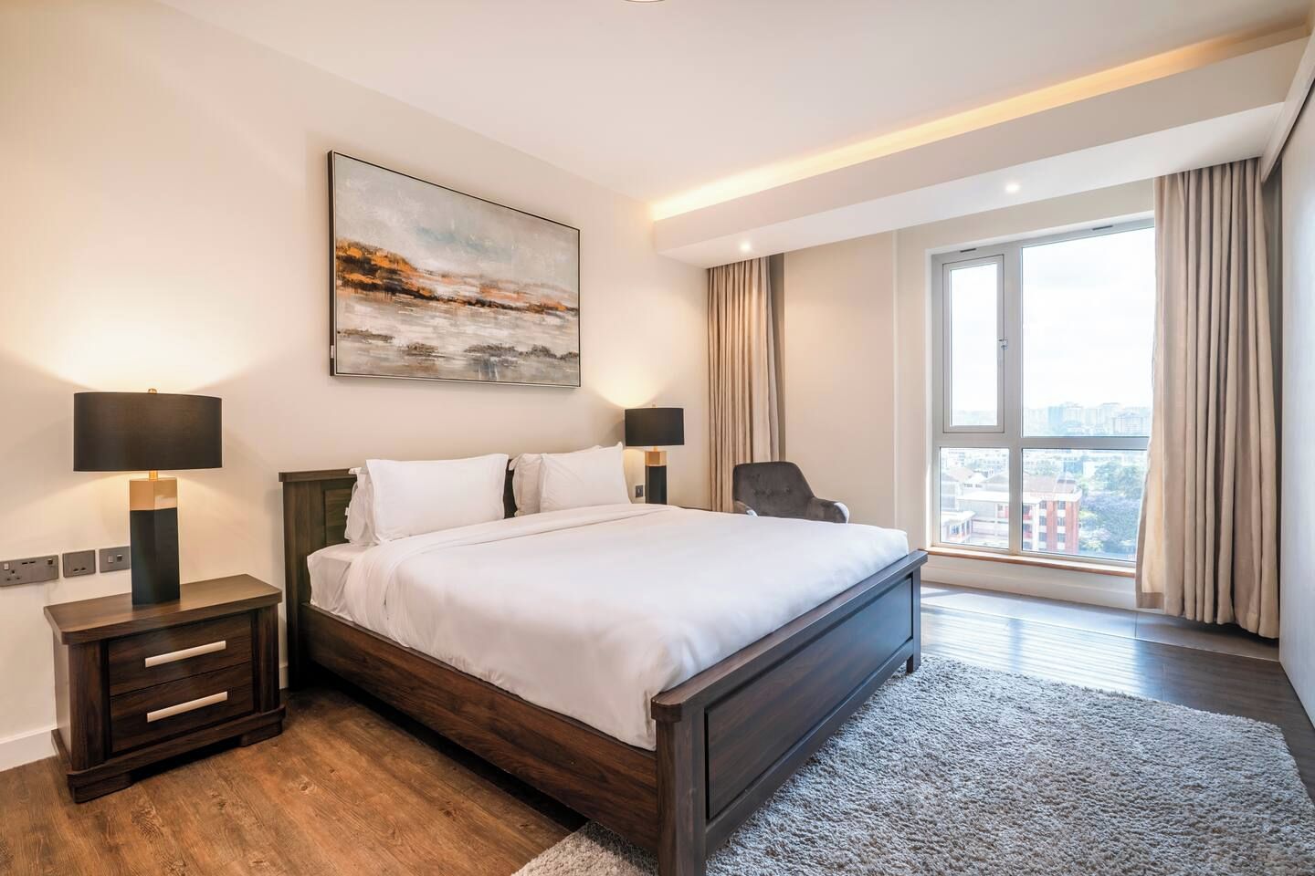 Une chambre d’hôtel moderne avec un grand lit, des lampes de chevet, un tableau, une vue à travers une grande fenêtre et un panorama sur la ville.