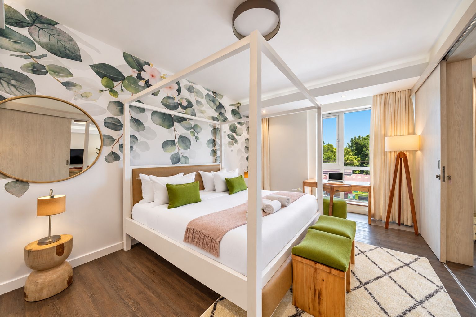 A modern hotel room with a four-poster bed, a large mirror, a wooden side table, and a view of the outdoors.