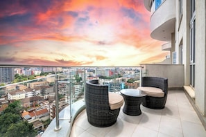 A balcony with wicker furniture, a glass railing, and a cityscape view during sunset. - Capital M Apartments by YourHost (Nairobi)