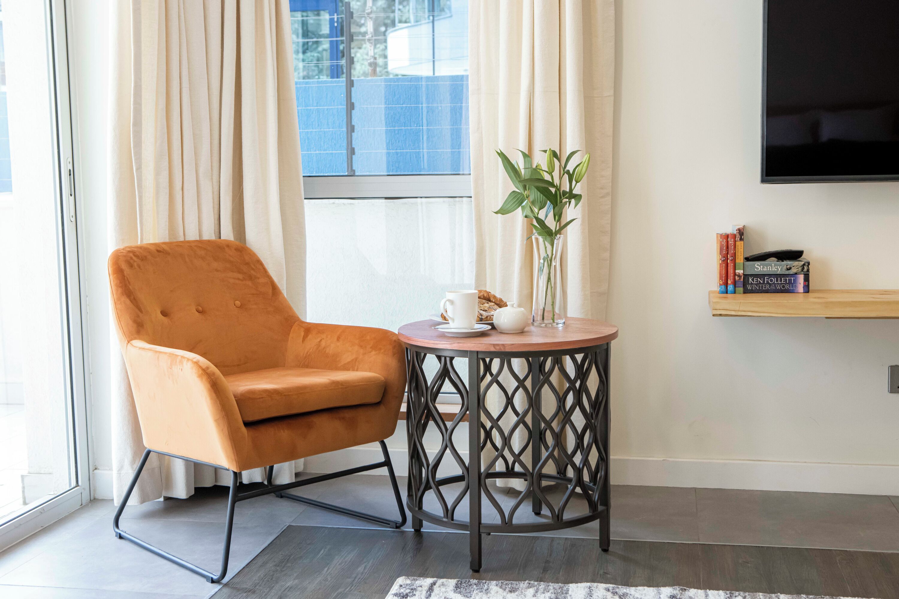 A modern living room with a comfortable orange armchair, a small round table with a plant, and a bookshelf with books.