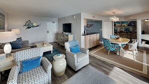 Una sala moderna con sofás estampados, una zona de comedor con sillas florales y una cocina con armarios de madera.