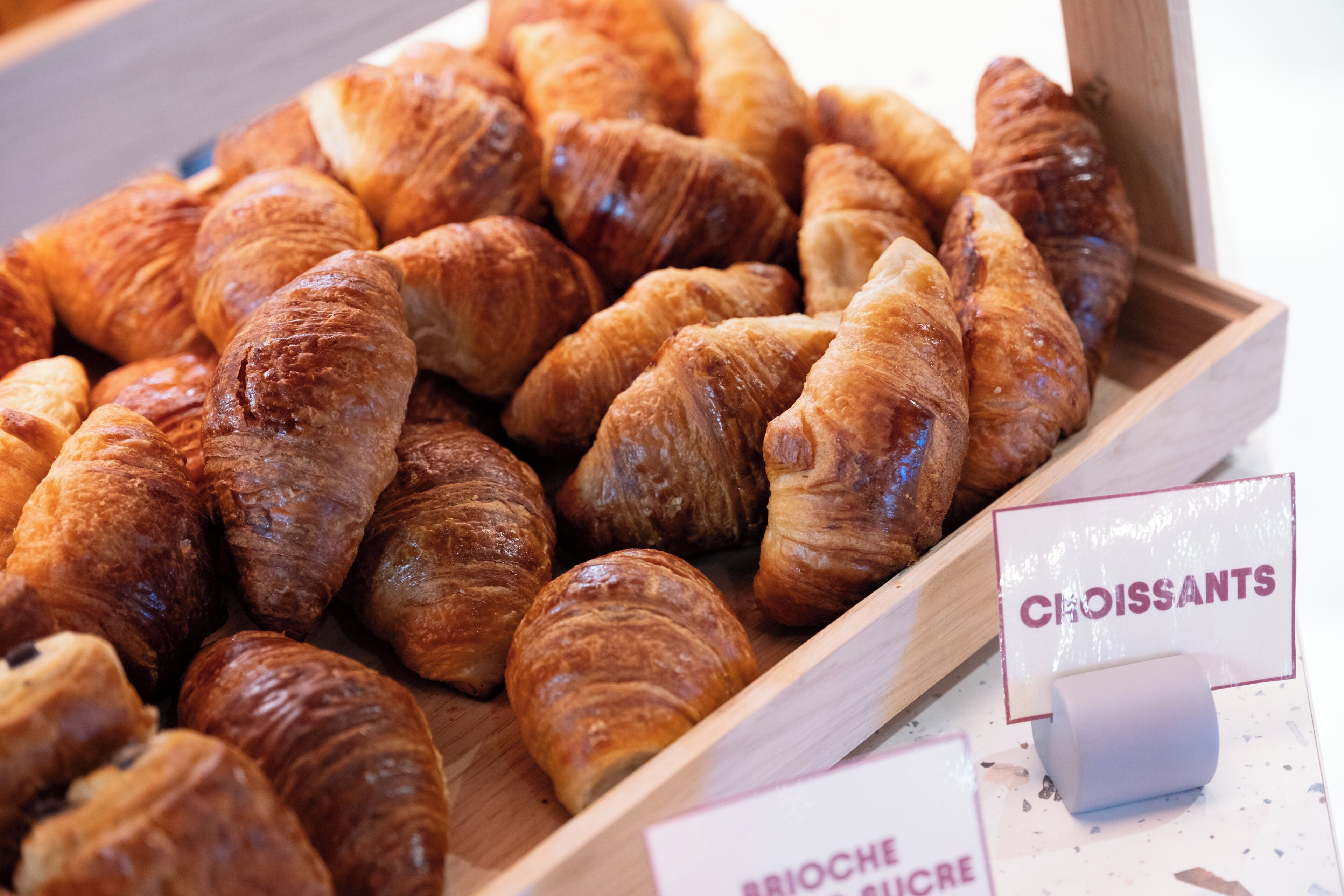A wooden tray filled with golden-brown croissants, labeled 'BRIOCHE SUGAR.'