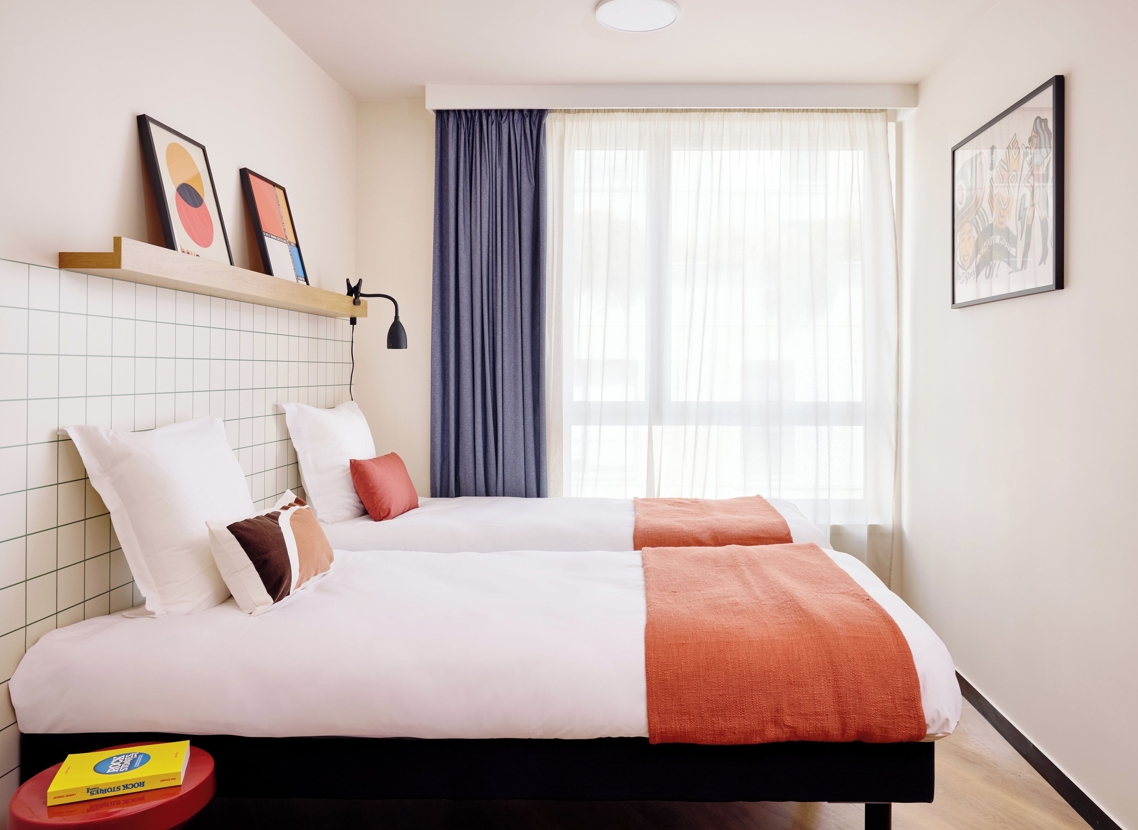 A hotel room with two beds, a window with sheer curtains, a shelf with framed artwork, and a small table with a book.