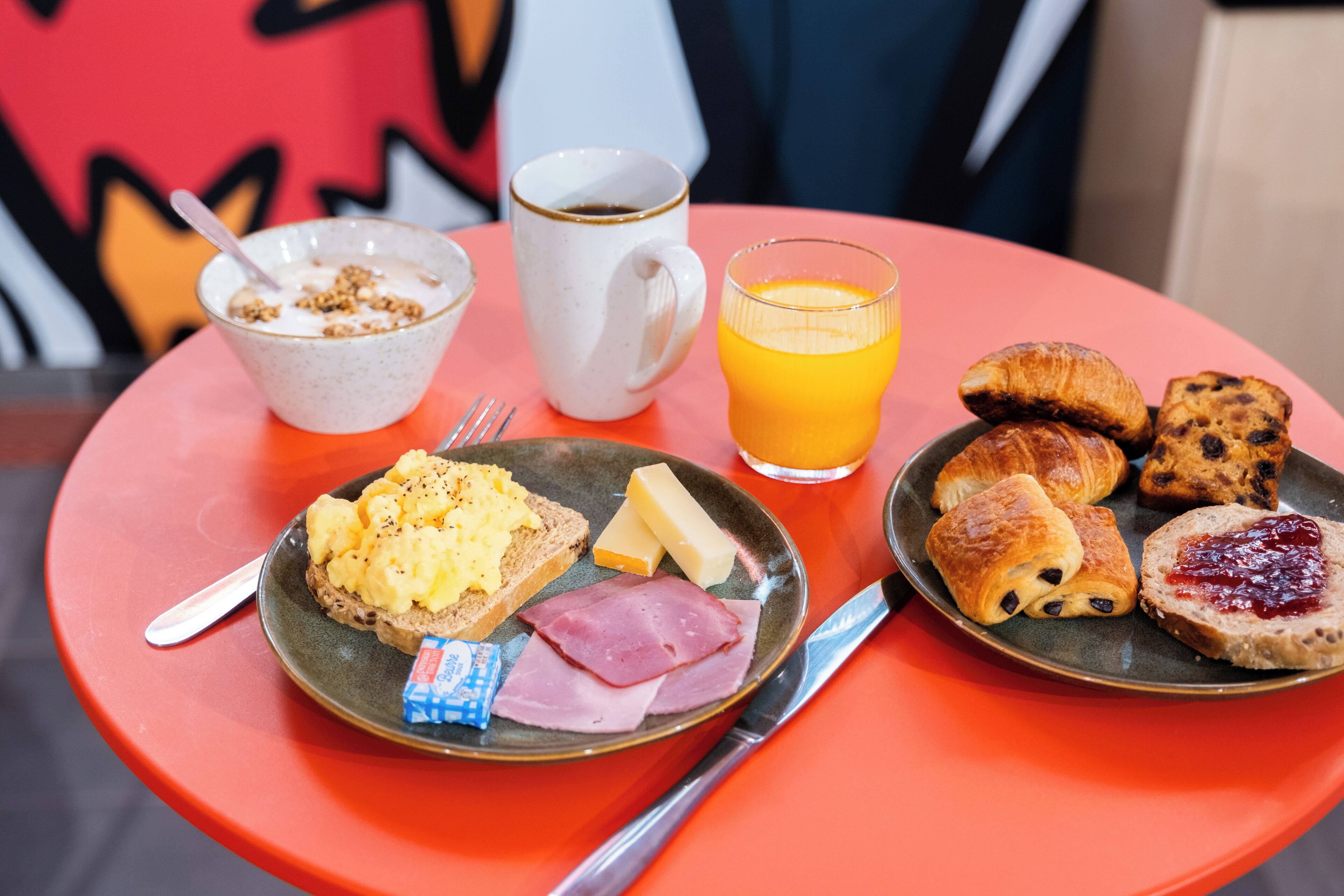 Ein Frühstückstisch mit Rührei, Käse, Schinken, Toast, einer Kaffeetasse, einem Glas Orangensaft und einer Auswahl an Gebäck.