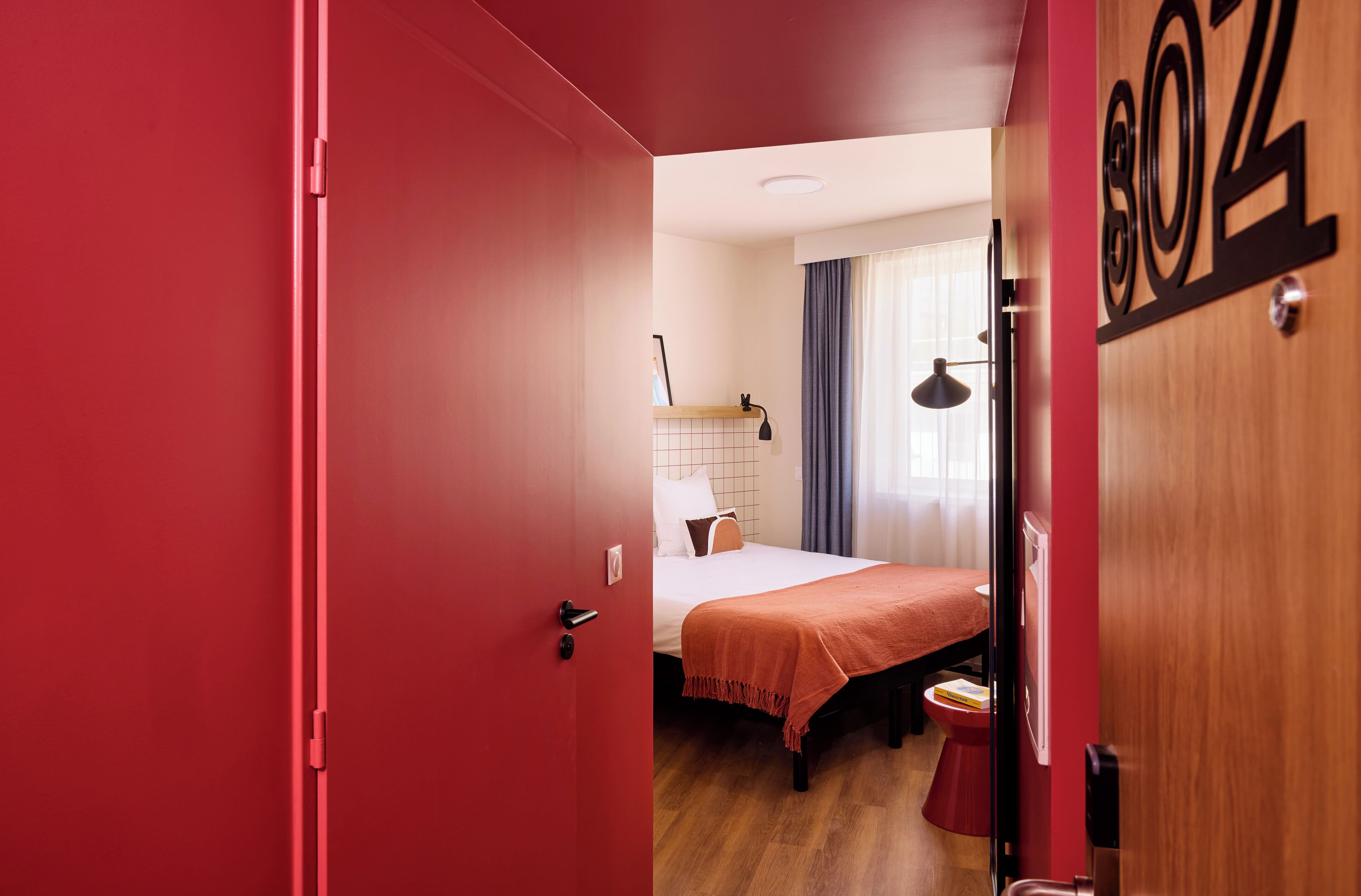 A hotel room with a red door, a bed with a white and orange bedspread, a wooden floor, and a window with curtains.