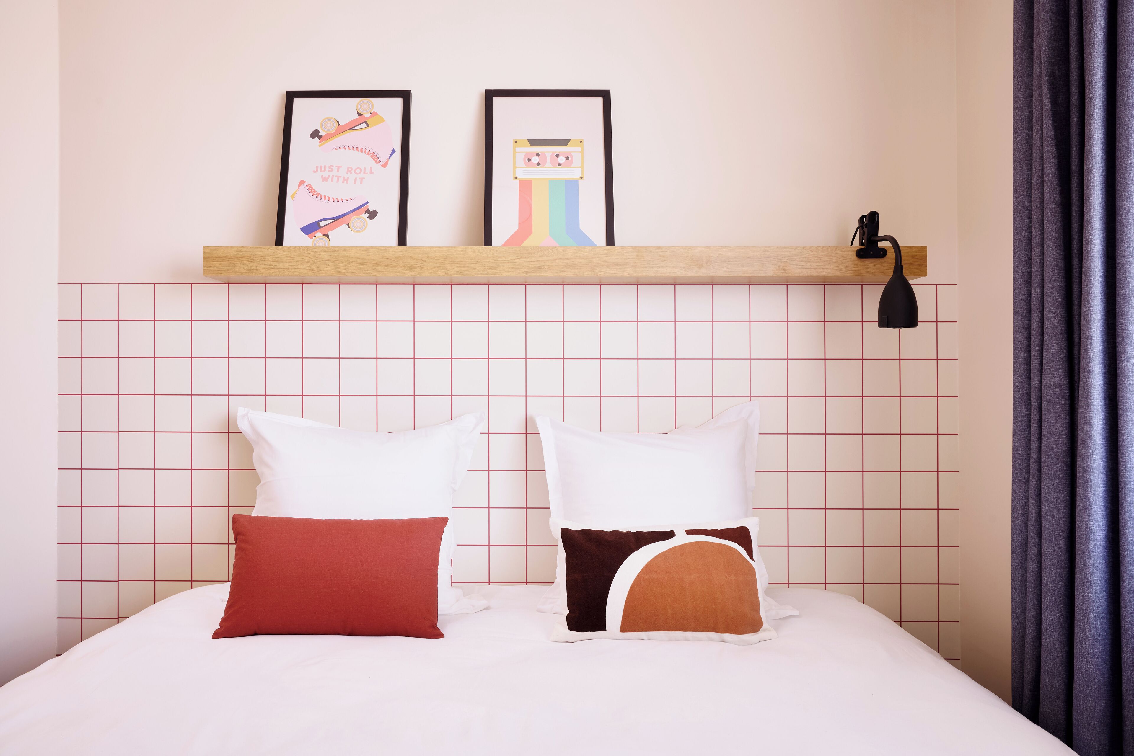 Una cama bien hecha con almohadas blancas y marrones, un estante con obras de arte enmarcadas arriba y una pared con diseño de cuadrícula.
